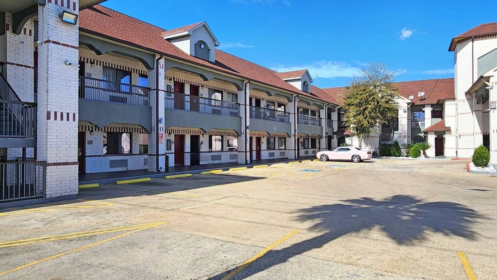 Two story building with covered stairs and walkways with exterior guest room entrances, parking spaces, landscaping with trees and shrubs