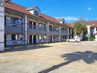 Two story building with covered stairs and walkways with exterior guest room entrances, parking spaces, landscaping with trees and shrubs