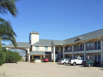 Hotel entrance with canopied drive through, two story building with exterior room entrances, covered walkways and stairs, parking spaces, landscaping with palm trees and bushes, parking spaces