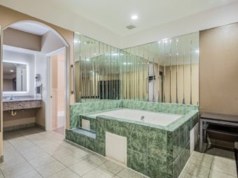 Jacuzzi with mirrored walls and tiled surround, tiled flooring, wall mounted full length mirror, night stand, alcove with vanity unit, wall mounted back lit mirror, wall mounted hairdryer and doorway to bathroom