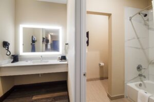 Vanity unit with coffee maker, hairdryer, ironging board with iron, doorway to bathroom with showertub and bathmat, tiled and laminte flooring