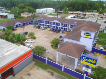 Hotel entrance with drive through canopy, brand signage, two story building with exterior walkways and room entrances, one story building with exterior room entrances, parking area, grassy areas with small trees