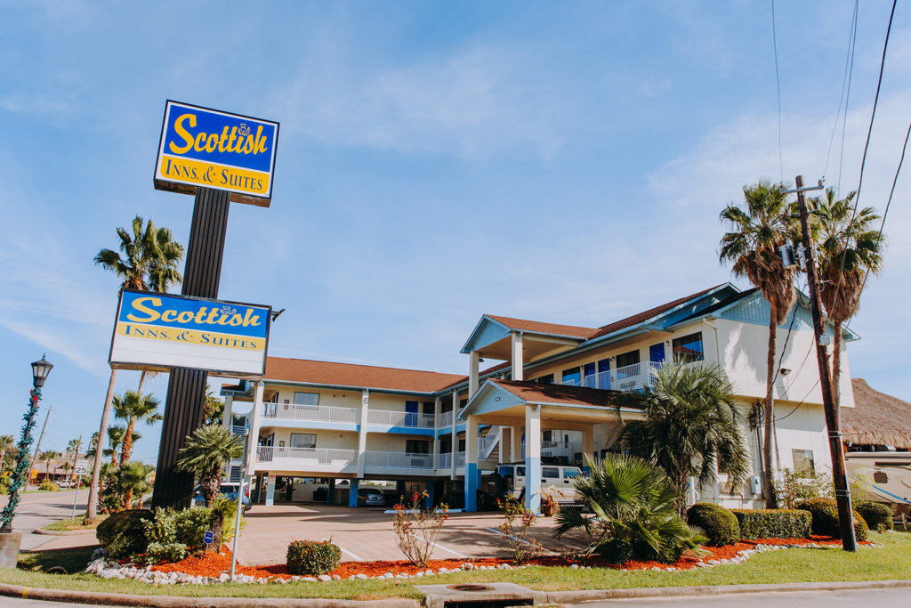 Hotel entrance, brand signage, two story building with exterior room entrances, covered walkways, stairs, covered parking, parking spaces, landscaping with grass, shrubs and trees