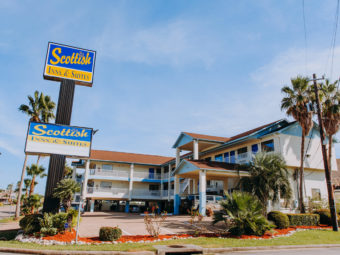 Hotel entrance, brand signage, two story building with exterior room entrances, covered walkways, stairs, covered parking, parking spaces, landscaping with grass, shrubs and trees