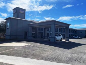 Hotel entrance with canopied drive through entrance, two story building with exterior guest room entrances and covered walkways. Parking spaces.