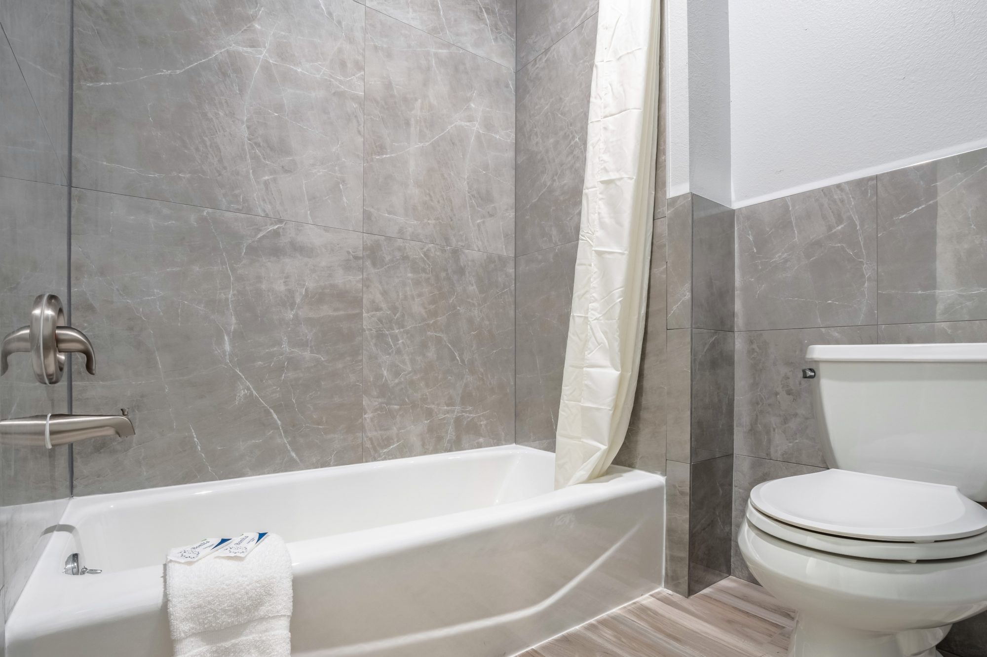 Shower tub with shower curtain, bathmat with bathroom amenities, toilet, laminated flooring