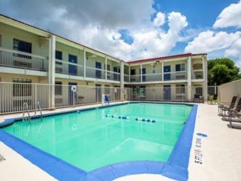 Exterior pool with safety fencing, concrete poolside with patio chairs, two story building with exterior room entrances and covered walkways