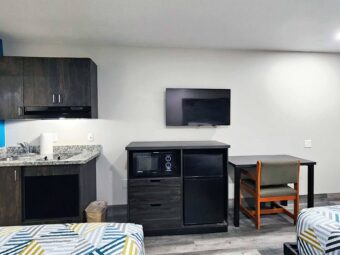 Two queen beds, wall cabinets with cooker hood, base cabinet with sink and hob, wall mounted TV, wooden unit with fridge and microwave, table and chair, laminate flooring