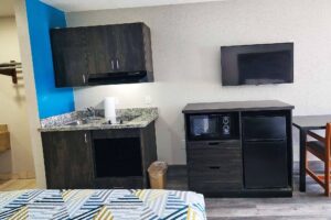 Wall cabinets with cooker hood, floor cabinet with sink and hob, wooden unit with fridge and microwave, wall mounted TV, table and chair, laminate flooring