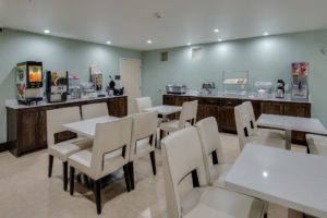 Tables and chairs, breakfast display counter with juice machine, coffee machine, waffle machine, toaster, breakfast pastry display cases, cereal dispensers, tiled flooring