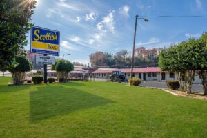 Brand signage, grassy area, small trees and bushes, single story building with exterior guest room entrances, parking spaces